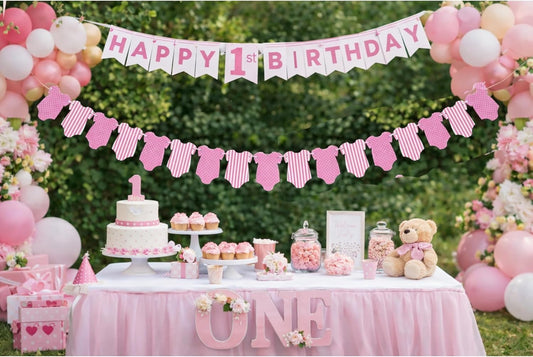 A pink and white baby shower themed first birthday party setup with a dessert table, cupcakes, a teddy bear, balloons, and a 'HAPPY 1st BIRTHDAY' banner against a green hedge background.