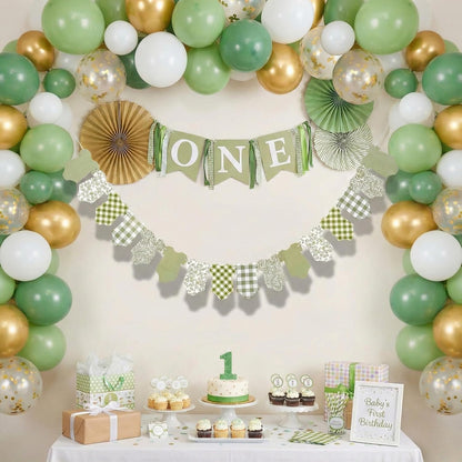 A green, white, and gold first birthday party decoration set up in a studio setting, featuring a balloon garland arch, a 'ONE' banner, paper fans, and a dessert table with cupcakes and gifts.