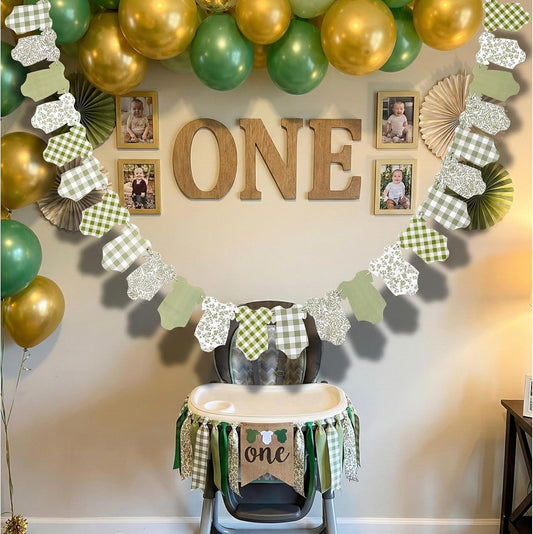A green, white, and gold first birthday party decoration set up in a nursery or playroom. The scene features a large arch of green and gold balloons, wooden 'ONE' lettering, and a decorative garland with green and white gingham checkered pieces, white lace-like pieces, and green leaf-shaped pieces.