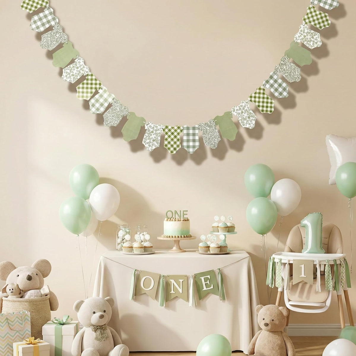 A styled baby first birthday party scene with a green and white gingham and solid green banner garland hanging on a beige wall. A decorated table below holds a small cake, cupcakes, balloons, a high chair, teddy bears, and a 'ONE' party banner.