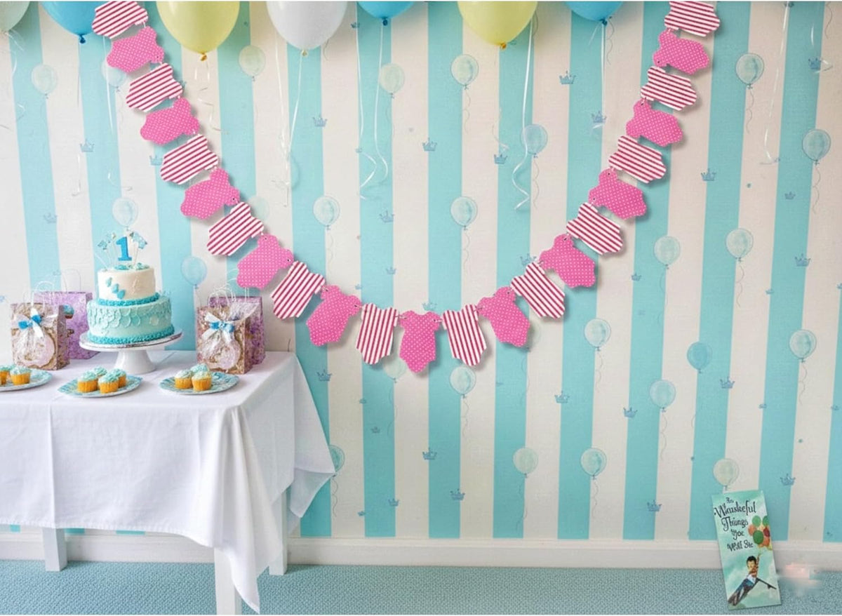 A pink and white striped heart-shaped bunting banner is displayed on a light blue and white striped party wall above a decorated baby shower or birthday table with a cake, balloons, and party supplies.