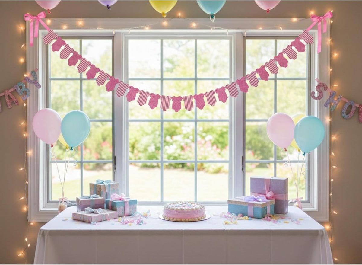 A decorated birthday party setup in front of a large window, featuring a pink pennant banner garland, pastel balloons, warm string lights, gift boxes, and a small cake on a white tablecloth.