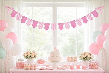 A styled baby shower dessert table decorated with pink and white baby clothes garland, pastel balloons, flowers, cupcakes, and a tiered cake in front of a bright window.
